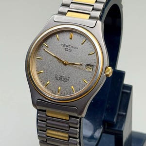 May include: A Certina DS wristwatch with a silver and gold-tone metal band and case. The watch face is gray with gold-tone hands and markers, and a date display at the 3 o'clock position. The watch is labeled "TITANIUM 100 METERS SAPPHIRE".