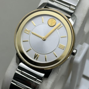 Könnte beinhalten: Silber- und goldfarbene Armbanduhr mit rundem Zifferblatt. Die Uhr hat ein silbernes Zifferblatt mit goldfarbenen Zeigern und Markierungen sowie eine goldfarbene Lünette. Das Armband besteht aus silberfarbenen Metallgliedern.
