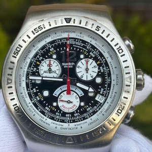 May include: A silver-toned Swatch watch with a black and white face. The watch features multiple dials, a red second hand, and a bezel marked with international city names. The watch band is also silver.