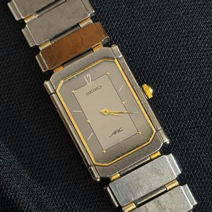 Pode incluir: Relógio de pulso Seiko Arc retangular com mostrador prateado e detalhes dourados. O relógio apresenta uma pulseira de metal prateado e dourado. O mostrador exibe o número 12 e as palavras "SEIKO" e "ARC".