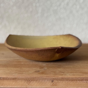 May include: A shallow, curved ceramic bowl with a speckled brown exterior and a smooth, matte yellow interior. The rim features a distinct, slightly indented notch on the right side, showcasing a natural, earthy texture and color contrast.