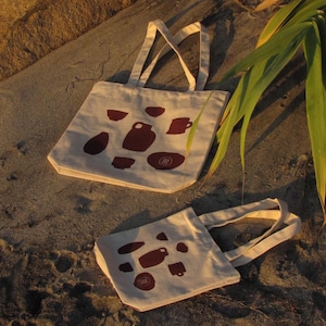 Puede incluir: Dos bolsas de mano beige con diseños de cerámica marrón se muestran sobre una superficie arenosa. Las bolsas tienen asas a juego y los diseños de cerámica incluyen tazas, cuencos y jarrones. Las bolsas están hechas de una tela resistente.