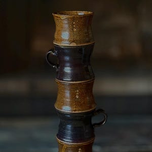 Pode incluir: Uma pilha de cinco canecas de cerâmica feitas à mão. As canecas alternam entre marrom escuro e marrom claro, com uma aparência texturizada e orgânica. Cada caneca tem uma pequena alça e uma borda ligeiramente alargada, criando uma estética rústica e artesanal.