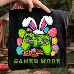 May include: Black square pillow with a green video game controller design, bunny ears, and Easter eggs. The pillow has the words "GAMER MODE" in green. Gold coins with the letters "H" and "B" are also visible.