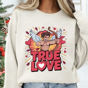 May include: Cream-colored sweatshirt featuring a cartoon Cupid with a bow and arrow, surrounded by hearts and arrows. The words "TRUE LOVE" are printed in red with a white heart. A festive design for Valentine's Day.