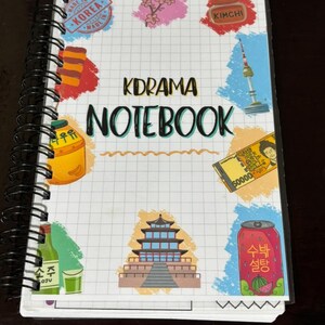Puede incluir: Un cuaderno con espiral con una cubierta blanca a cuadros. La cubierta presenta el texto "KDRAMA NOTEBOOK" en negro, junto con ilustraciones coloridas de elementos culturales coreanos, incluyendo comida, monumentos y moneda. El cuaderno está abierto.