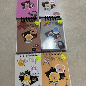 May include: A selection of spiral-bound notebooks with illustrated covers. Each notebook features cartoon character stickers and handwritten text. The notebooks are in various colours, including pink, brown, and white.