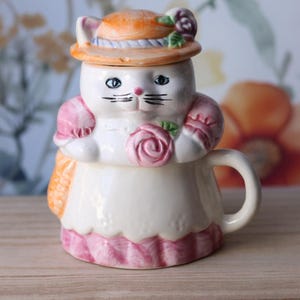 May include: A ceramic cat-shaped teacup with a lid, featuring a white body, pink and orange accents, and a hat. The cat has black whiskers and blue eyes. A pink rose decorates the front. The teacup has a handle.