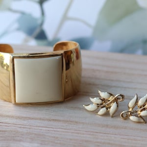 May include: Gold-tone cuff bracelet with a cream-colored square center and matching leaf-shaped earrings. The earrings and bracelet are set on a light-colored wooden surface.