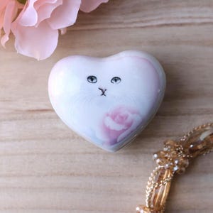 May include: A heart-shaped trinket box with a white and pink cat face design. The box features a rose illustration and is accented with gold-colored jewelry and a pink flower.