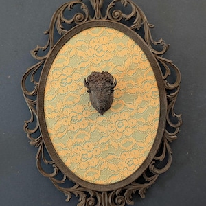 May include: An oval, dark brown framed wall hanging with a decorative floral design. The center features a patterned fabric in yellow and green, with a dark brown bison head sculpture mounted in the center. The frame has ornate detailing.