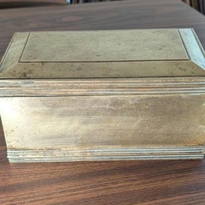 May include: A rectangular brass box with a hinged lid. The box has a simple, elegant design with a slightly aged appearance. The lid and sides are adorned with subtle linear details. The box is likely used for storing small items.