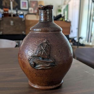 May include: A brown, antique-style ceramic bottle with a rounded body and a narrow neck. The bottle features a raised emblem on the front. The emblem appears to be a religious symbol. The bottle is likely used for decorative purposes.