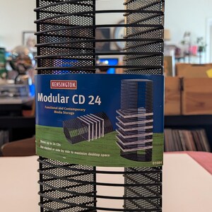 May include: Black metal CD storage rack with a curved front and a mesh design. The product label reads "Modular CD 24" and "Functional and Contemporary Media Storage". It can store up to 24 CDs.