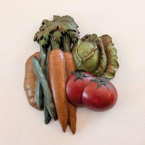 May include: A wooden wall hanging featuring a collection of carved vegetables. The arrangement includes carrots, tomatoes, a cabbage, and other vegetables, painted in shades of brown, orange, green, and red. The piece is designed to resemble a still life.