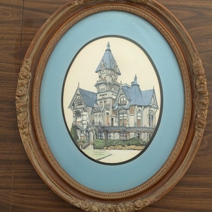 May include: An oval framed print of a Victorian house with a blue roof and a light blue background. The frame is a dark wood color with ornate detailing. The print is centered within the frame.
