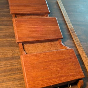 May include: A brown wooden wall-mounted organizer featuring three tiered pockets for storage and three metal hooks at the bottom. A wooden ruler is placed alongside the organizer, showing a length of approximately 18 inches for the unit.