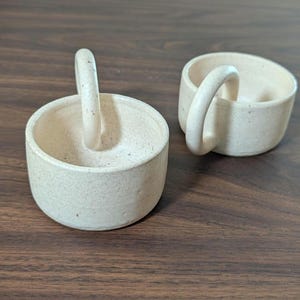 May include: Two small, off-white ceramic bowls with arched handles. The bowls have a speckled texture and a cylindrical shape. The handles are rounded and attached to the sides of the bowls. The bowls are sitting on a dark wooden surface.