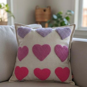 Peut inclure: Coussin carré avec un fond crème et neuf cœurs brodés. Les cœurs sont dans des tons de rose et de violet. Le coussin est posé sur un canapé clair.
