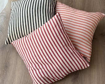 May include: Three square throw pillows with striped patterns. One pillow has black and white stripes, another has red and white stripes, and the third has peach and white stripes. The pillows are arranged on a wooden surface.