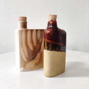 May include: Two ceramic bottles with cork stoppers. One is a light beige with brown streaks, the other is brown and beige. The bottles have a rectangular shape with rounded edges, and are likely for decorative use.