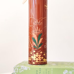 Può includere: Un contenitore decorativo alto, cilindrico, marrone rossiccio con un design floreale e geometrico in oro e verde. Il contenitore ha un coperchio con un nastro bianco attaccato. Il contenitore è appoggiato su una pila di libri.