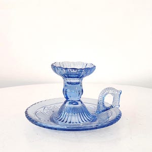 Puede incluir: Un candelabro de vidrio azul claro con base redonda y asa. El candelabro tiene un diseño acanalado y un borde festoneado. El asa es arqueada y tiene un diseño decorativo. El candelabro está sobre una superficie blanca.