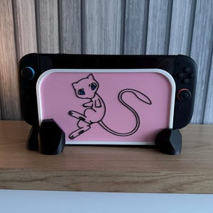 Può includere: Supporto nero per console di gioco con un inserto rosa raffigurante il personaggio dei cartoni animati Mew. Il supporto ha una base nera ed è progettato per contenere una console di gioco. La console ha pulsanti blu e rossi.