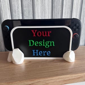 Può includere: Una console di gioco nera tenuta da un supporto bianco a forma di roccia geometrica. Il supporto presenta un pannello rettangolare nero con il testo "Your Design Here" in lettere in rilievo rosse, verdi e blu. La console ha controller neri con accenti blu e rossi.