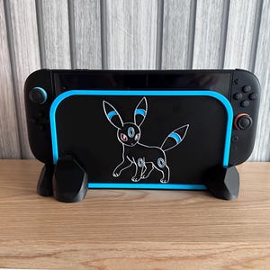 May include: Black gaming console stand with a blue border and an Umbreon cartoon design. The stand is made of black plastic and has a matching black base. The console is resting on the stand.