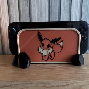 May include: Black Nintendo Switch console with a custom Eevee-themed skin and a matching stand. The skin features a cartoon Eevee character on an orange background. The stand is black and holds the console upright.