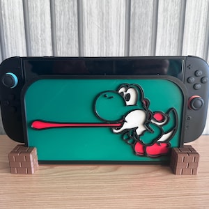 May include: A black Nintendo Switch console sits in a green holder with a Yoshi character design. The holder is supported by two brown brick-like stands. The Yoshi design has white, red, and black accents. The background is a light wood surface and a vertical wood-slat wall.