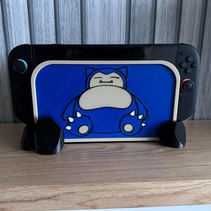 May include: A black Nintendo Switch console with a blue and white Snorlax-themed stand. The stand features a blue background with a cartoon image of Snorlax. The console is resting on a wooden surface.