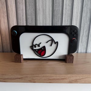 May include: Black and white Nintendo Switch console with a white center panel featuring a cartoon ghost design. The console rests on two brown, cube-shaped stands, set against a wooden surface and a gray-toned background.