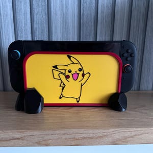 May include: Black Nintendo Switch console with a yellow screen featuring a Pikachu graphic. The console is supported by two black stands. The screen has a red border. The background is a light blue and gray vertical striped wall.