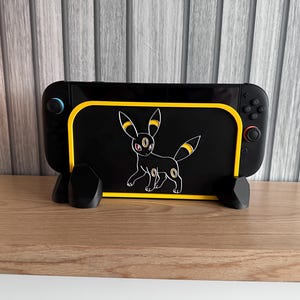 May include: Black gaming console stand with a yellow border and a white outline drawing of an Umbreon Pokémon. The stand has a black base and is designed to hold a gaming console. The console has blue and red buttons.