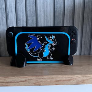 May include: Black gaming console stand with a blue and black graphic of a dragon. The stand is on a wooden surface. The dragon has blue wings and accents. The stand is designed to hold a gaming console.