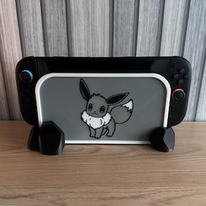 Puede incluir: Soporte negro y gris para Nintendo Switch con un gráfico de Eevee. El soporte tiene un borde blanco y está sostenido por tres formas geométricas negras. Diseñado para sostener una consola Nintendo Switch.