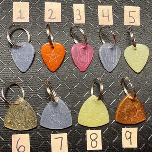 Puede incluir: Una colección de nueve llaveros en varias formas y colores. Cada llavero presenta un anillo de metal y un dije decorativo con diseños grabados, incluyendo estrellas, símbolos y un sol. Los dijes son en tonos azules, naranjas, rosas y amarillos.