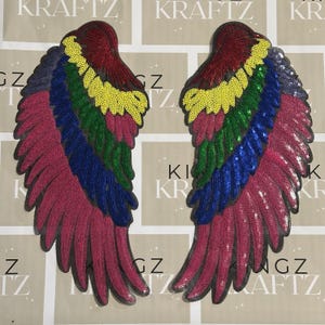 May include: Two sequined wing patches in a rainbow of colours. The wings are arranged symmetrically, with each wing featuring a gradient of colours including red, yellow, green, blue, and purple. The patches are likely for decorative purposes.