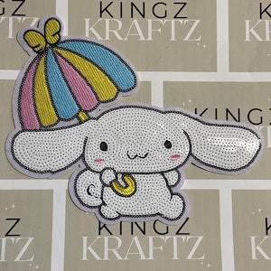 May include: A white sequined patch of a cartoon character with long ears, holding a yellow umbrella with blue, pink, and yellow stripes. The patch has a black border and is on a light background with the text "KINGZ KRAFTZ".
