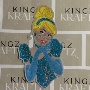 May include: Embroidered patch depicting a cartoon Cinderella. The patch features a yellow bun, blue dress, and gloves. The background shows the text "KING KRAFT".