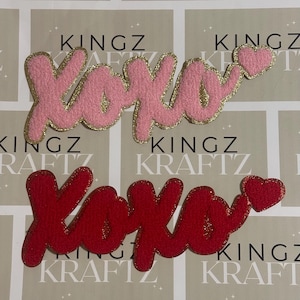 Peut inclure: Deux écussons brodés avec le mot "XOXO" en écriture cursive. L'un est rose avec une bordure dorée pailletée et un cœur. L'autre est rouge avec une bordure dorée pailletée et un cœur.