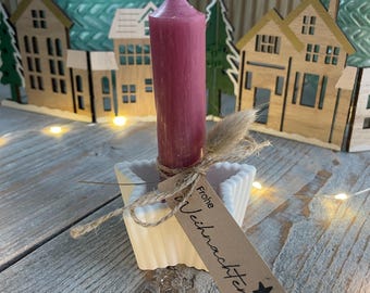 May include: A burgundy candle in a white square holder, tied with twine and a small tag. The tag has text and a star. Wooden house and tree decorations are in the background, on a wooden surface.