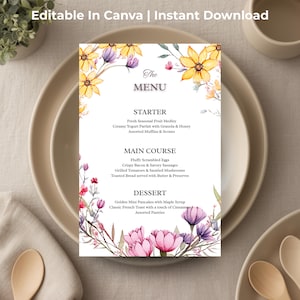 May include: A menu card with floral decorations and the text "The MENU". The card features a list of dishes and is adorned with watercolour flowers in yellow, pink, and purple. The menu is on a white background, surrounded by plates, cups, and wooden spoons.