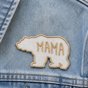 May include: A light blue denim jacket with a white bear patch that says "MAMA" in tan letters. The patch is sewn onto the jacket's chest area. The jacket has a buttoned pocket and a button closure.