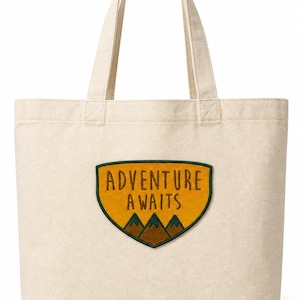 May include: A cream-colored tote bag with a patch that reads "ADVENTURE AWAITS." The patch features a yellow background with a green border and brown mountain illustrations. The bag has two handles.