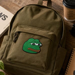 Peut inclure: Un sac à dos en toile vert olive avec un écusson brodé centré représentant un personnage de grenouille verte aux grands yeux et à la bouche brune. Le sac à dos possède une poche avant zippée, un compartiment principal et une poignée supérieure.