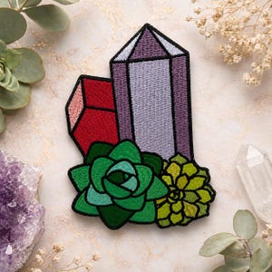 Op de afbeelding: Geborduurde patch met een paarse en grijze kristal, een rode kristal en twee vetplanten in groen- en geeltinten. De patch heeft een zwarte rand en is bedoeld voor kleding of accessoires.