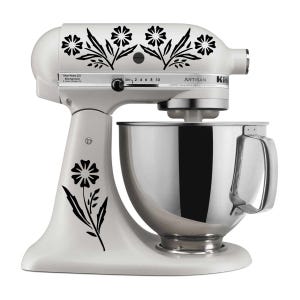 Puede incluir: Una batidora de pie KitchenAid Artisan blanca con un cuenco de acero inoxidable. La batidora está decorada con calcomanías florales negras. La batidora tiene un dial de control de velocidad y el logotipo de KitchenAid es visible en el lateral.